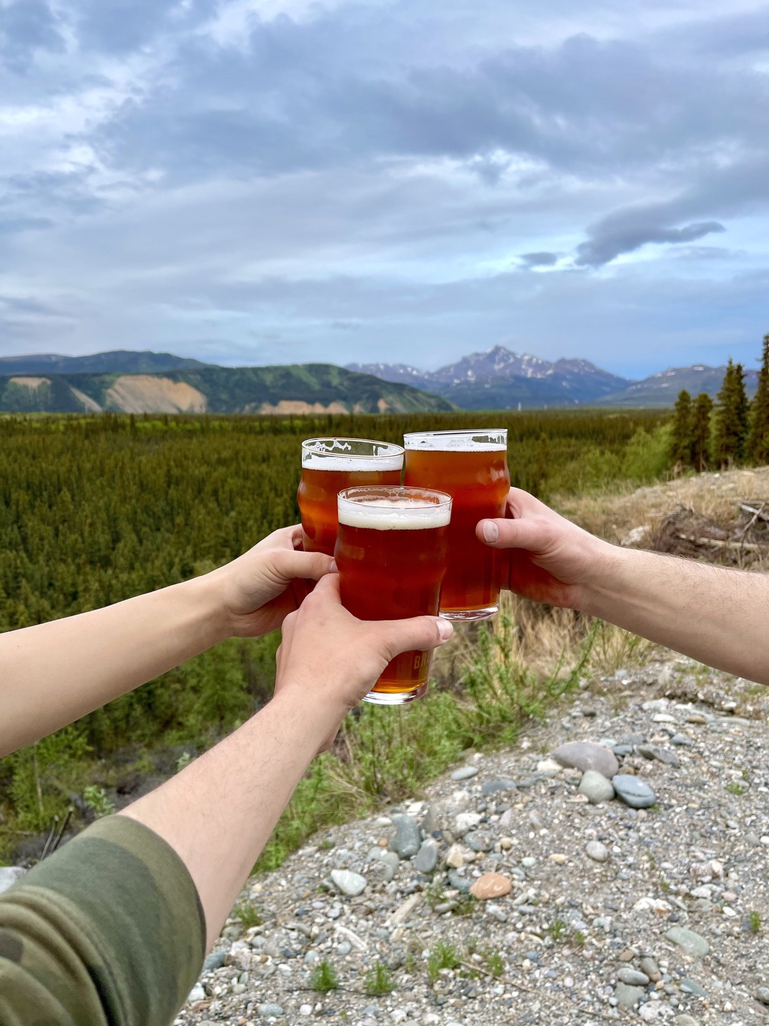 Three beers raised in a toast against an Alaska mountain landscape